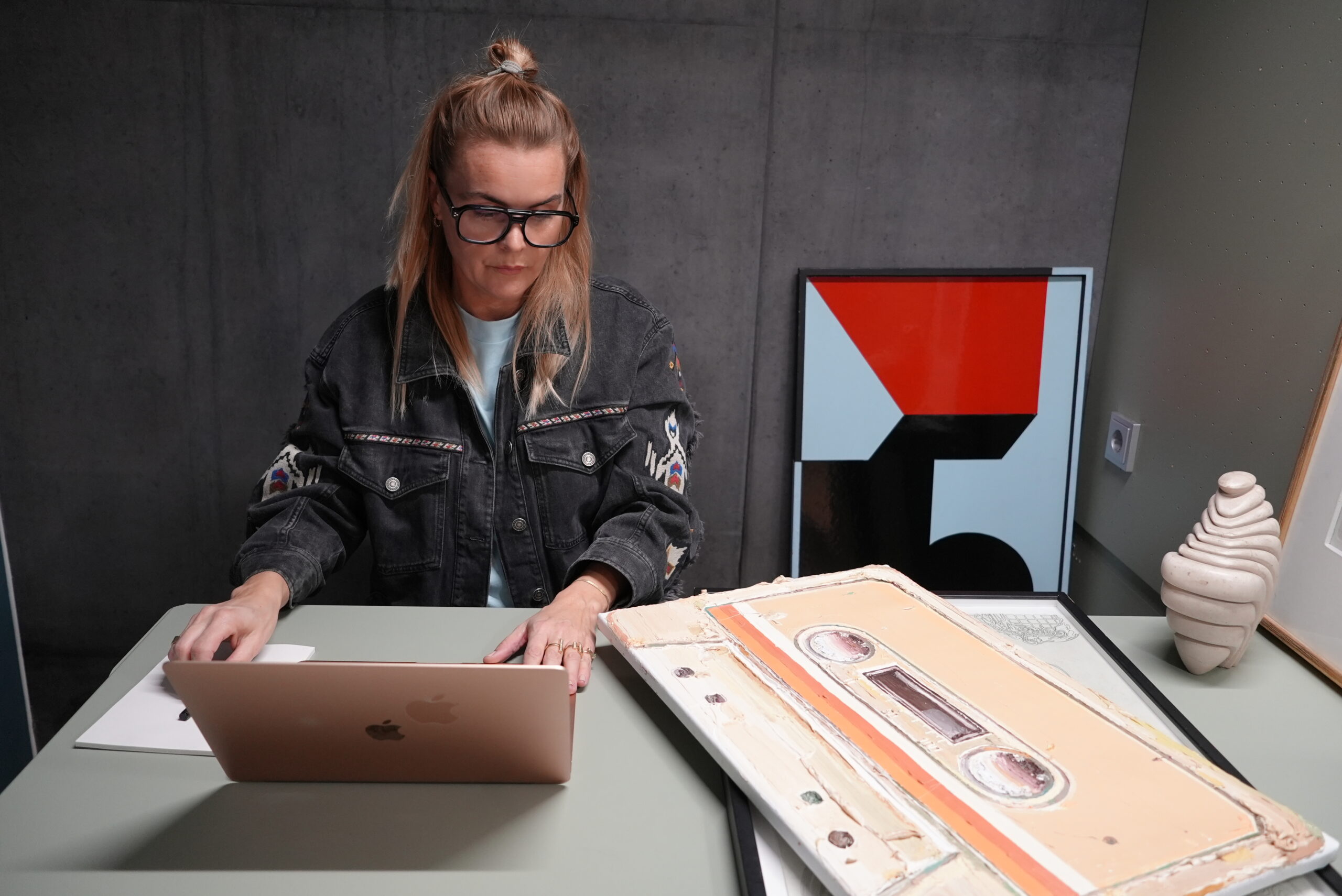 Annemette Müller arbejder ved computer med værker af Carl Beier, Jesper Neergaard og Søren Bjørn i baggrunden.